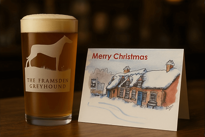 The Framsden Greyhound official pint glass sits next to a hand-painted Framsden Greyhound Christmas card atop a wooden table.